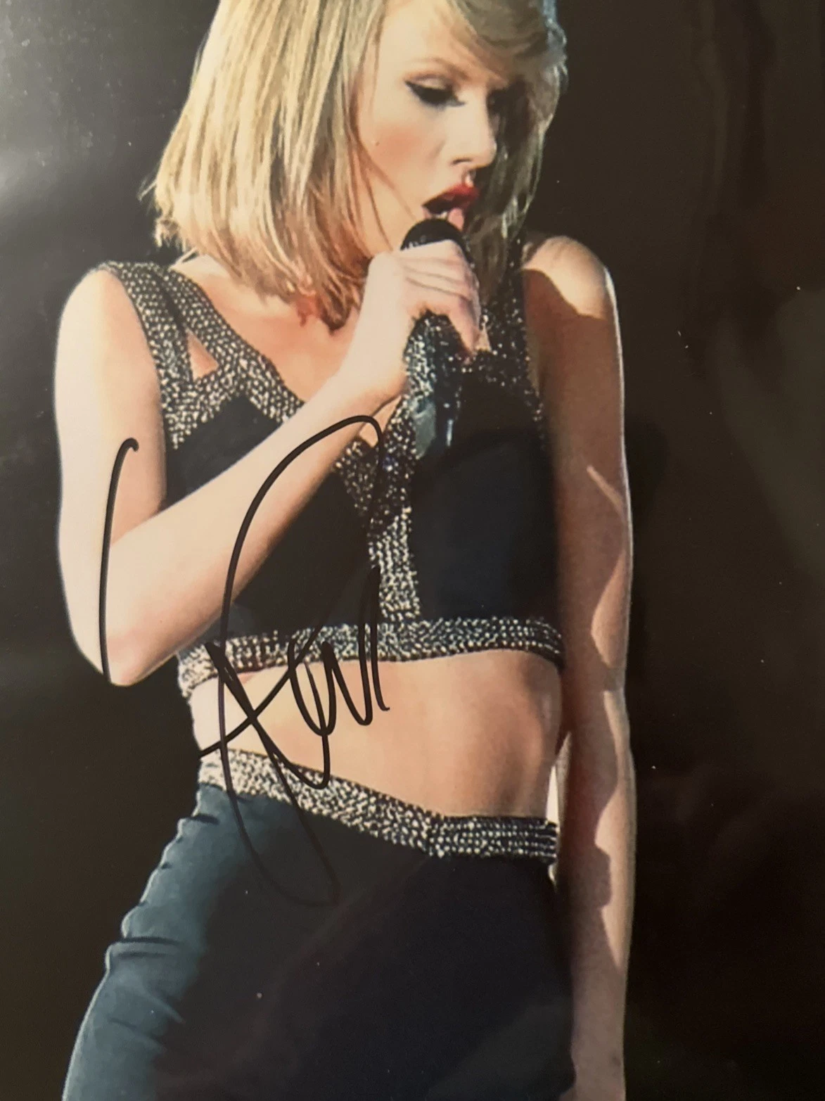 A colour concert photograph of Taylor Swift singing into a handheld microphone, signed in broad black marker straight across the centre of the image.
