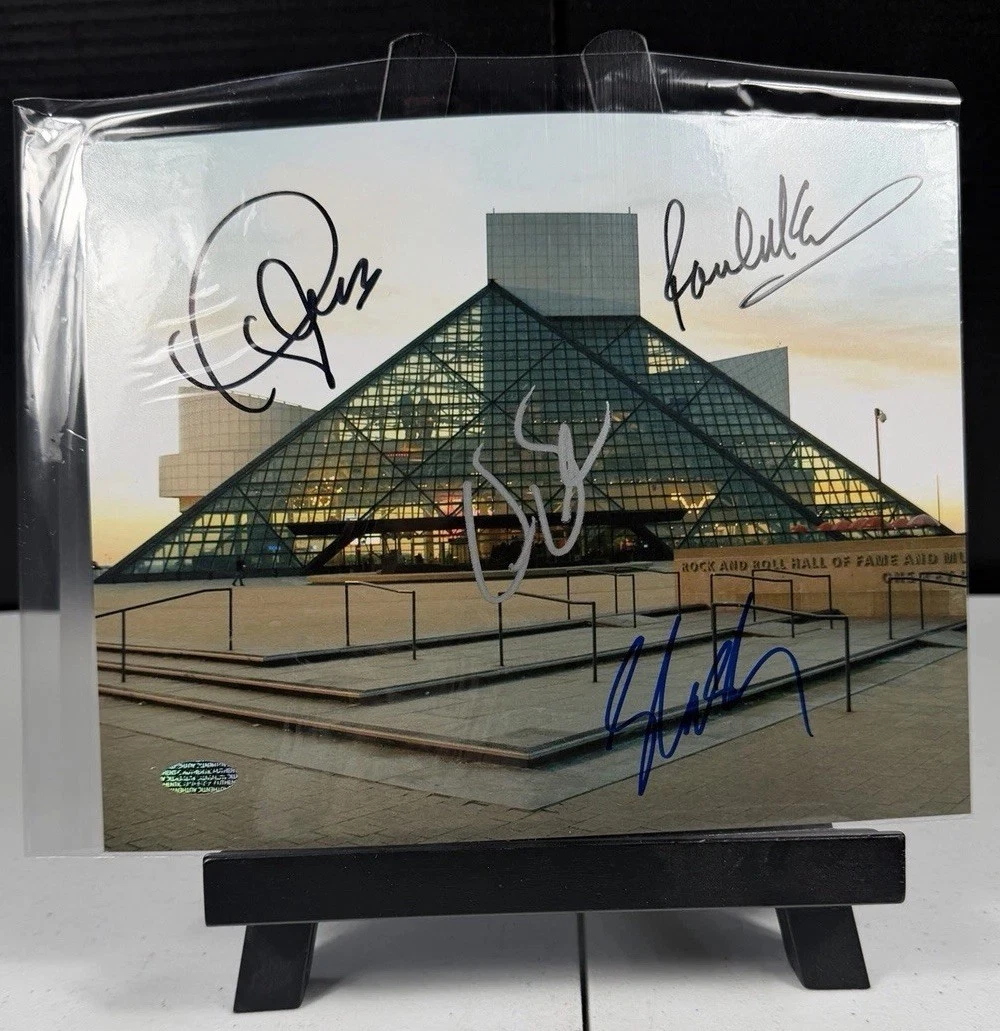 A colour photograph of the Rock and Roll Hall of Fame building, sleeved on a display stand, signed across the sky and the plaza by four different hands in black, silver, and blue.