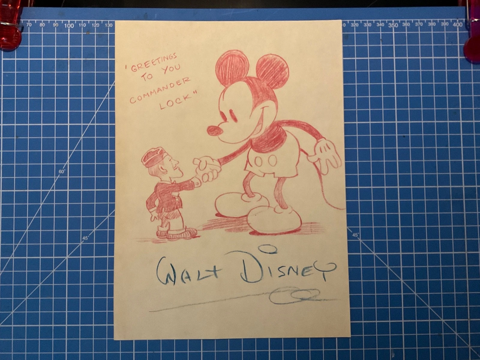 A red pencil drawing of Mickey Mouse shaking hands with a uniformed serviceman, captioned "Greetings to you Commander Lock", signed Walt Disney in blue underneath.